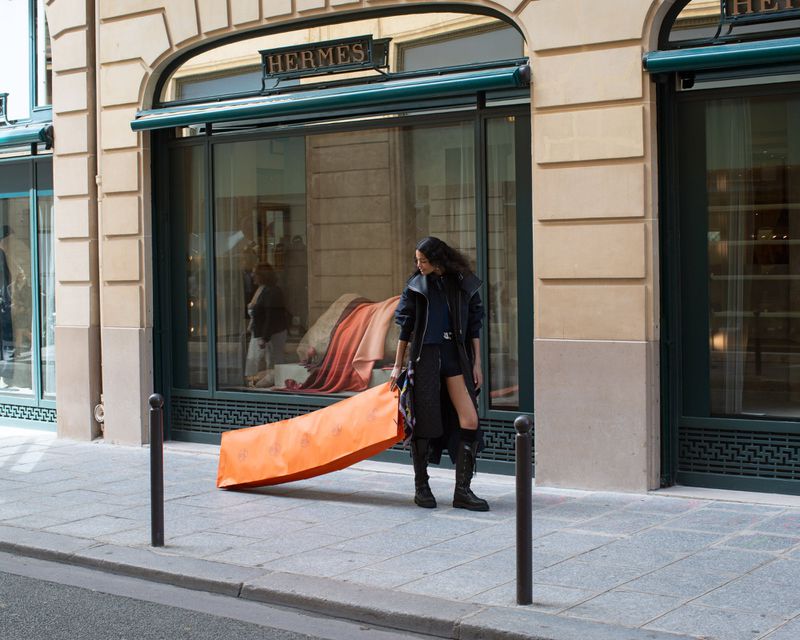 Woman in black coat walks past an Hermes store holding a large orange package in an urban setting, with a sidewalk and beige building.