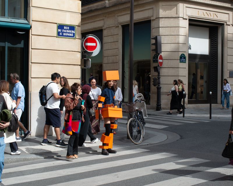 Pedestrians cross a Paris street at a crosswalk, including one person in a costume of orange rectangular blocks.