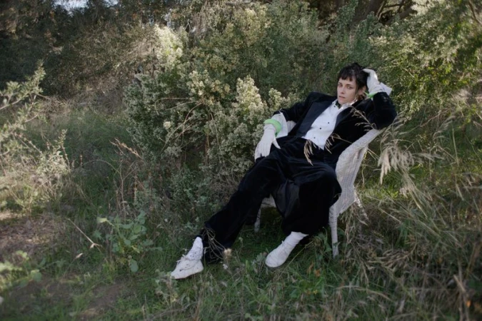 Adult in a black tuxedo suit and white gloves lounges in a wicker chair in a grassy, wooded environment with green foliage and trees.