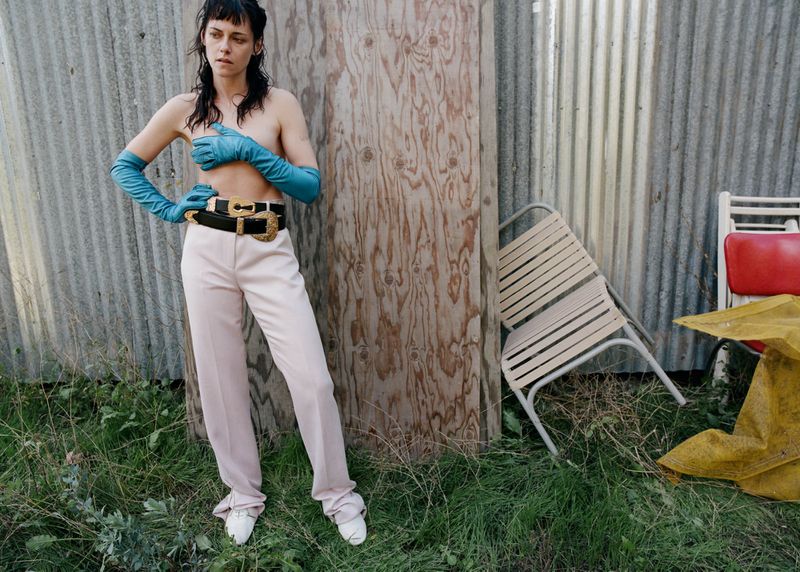 Fashionable woman in pink pants, gloves posing outdoors against a weathered wall with chairs, creating a stylized portrait.