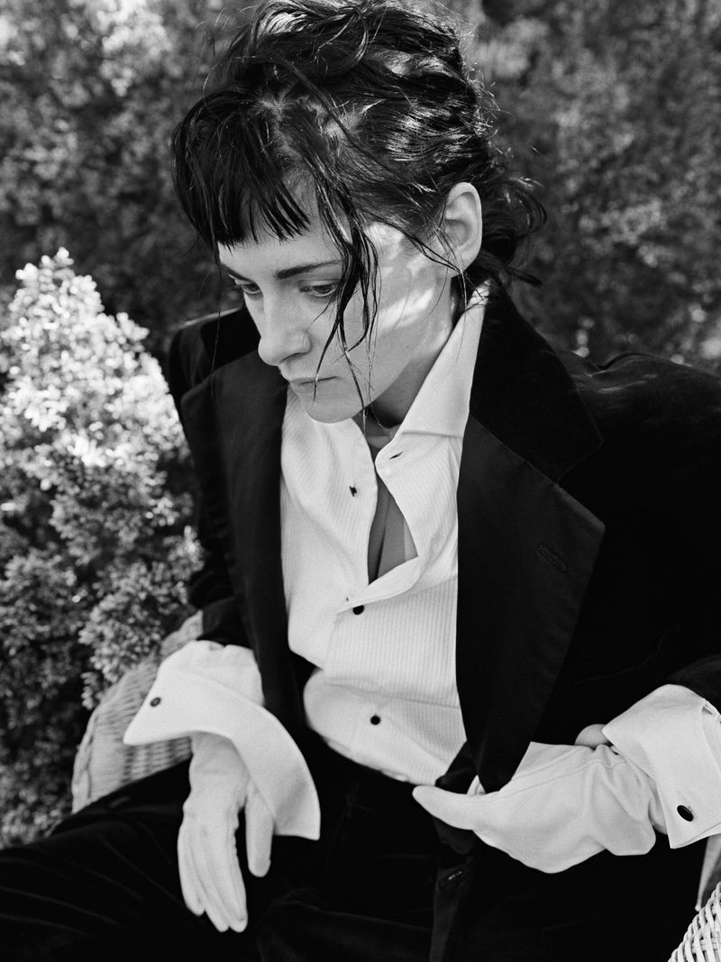 Monochrome portrait of a seated adult woman wearing a tuxedo and gloves, outside with leafy green background, looking downwards.