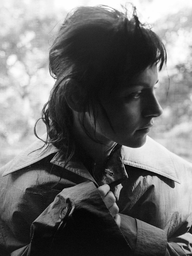 Black and white portrait of a woman with dark hair wearing a buttoned trench coat, gazing thoughtfully near a window.