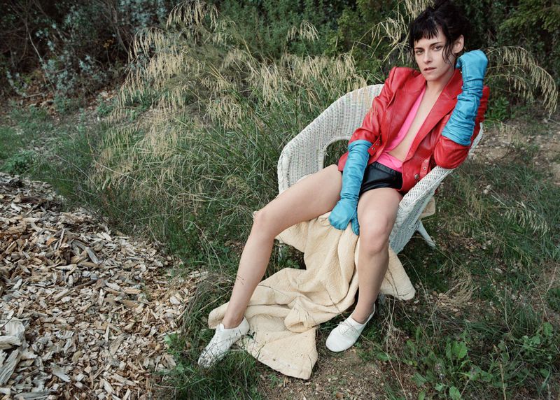 Woman in red jacket and blue gloves sits in wicker chair surrounded by greenery, wearing white shoes, creating a fashion-forward image.