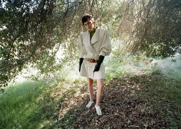 Kristen Stewart stands outdoors in an oversized cream belted coat dress with long black leather gloves among trees.