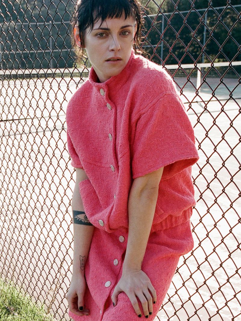 Portrait of a woman in a pink button-down top and skirt standing outdoors in front of a chain-link fence on a sunny day.