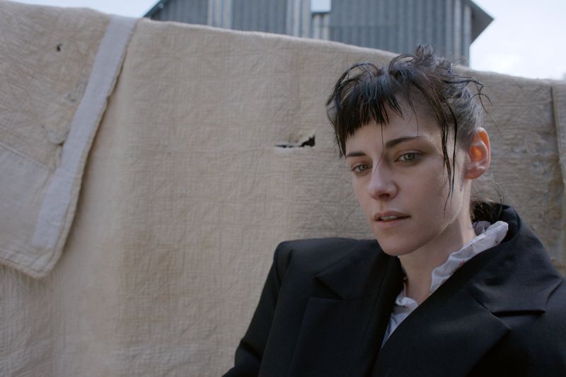 Portrait of a white adult woman in a black jacket standing in front of a neutral-colored canvas and a corrugated metal structure outdoors.