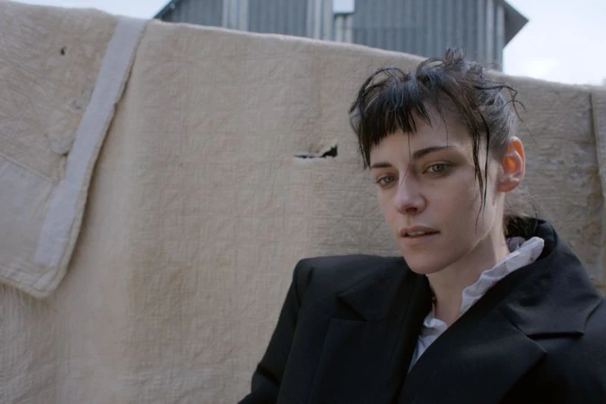 Portrait of a white adult woman in a black jacket standing in front of a neutral-colored canvas and a corrugated metal structure outdoors.