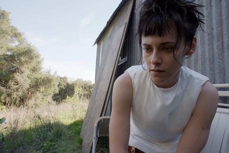 Portrait of a fair-skinned adult female with dark hair, bangs, and a white tank top sitting outdoors near a weathered shed and green foliage.