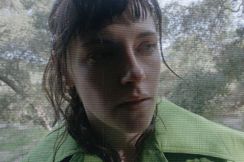 Close-up of an adult woman with dark hair and light eyes, wearing a collared green shirt, posed indoors with greenery visible outside.