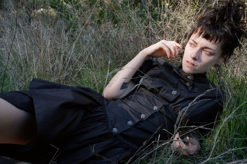 A woman lies in tall grass wearing a dark dress; she looks off into the distance with her hand raised near her face outdoors.