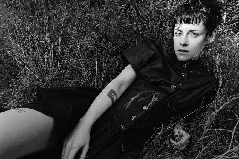 Monochrome portrait of a woman lying in tall grass, wearing a dark dress with decorative buttons, and showcasing arm and leg tattoos.