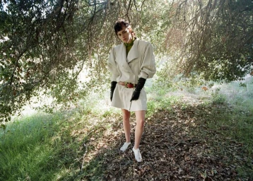 Kristen Stewart stands outdoors in an oversized cream belted coat dress with long black leather gloves among trees.