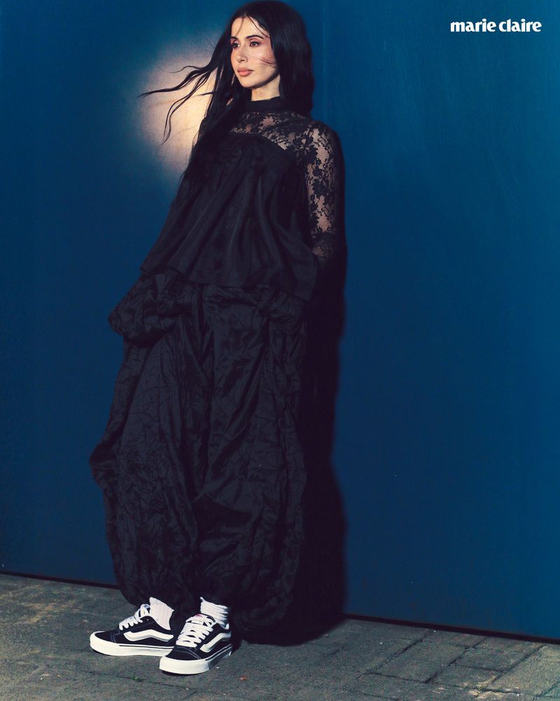 Full-length portrait of a woman in a black lace dress and sneakers standing against a blue wall, fashion editorial, Marie Claire logo visible.