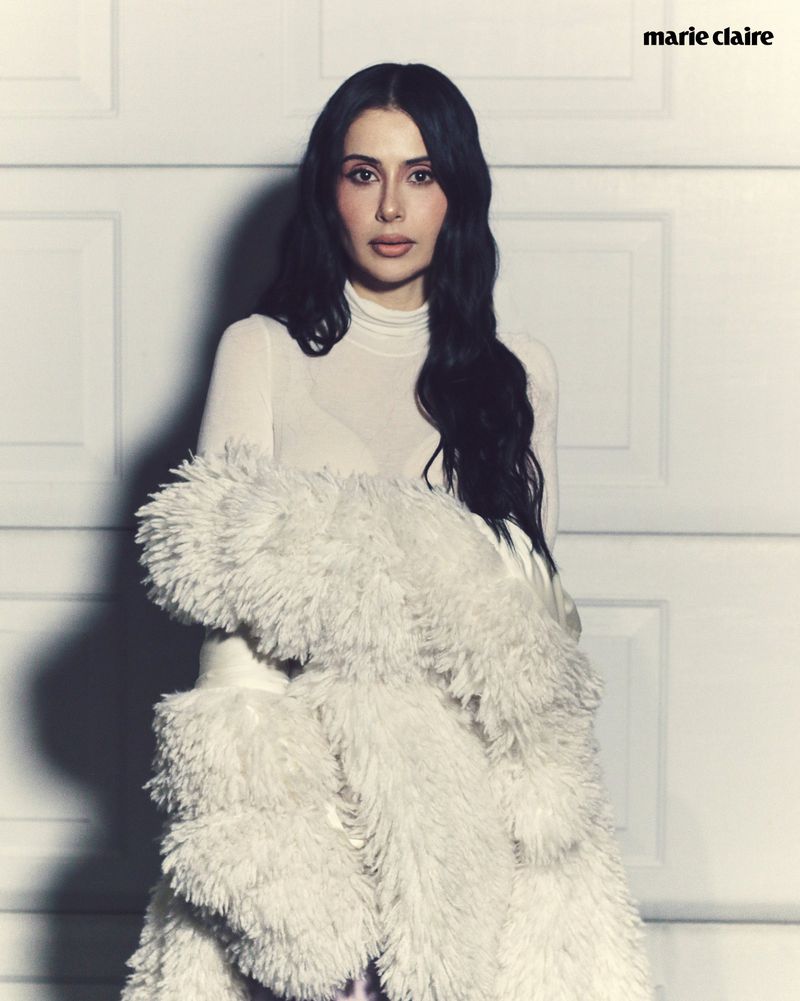 Portrait of a woman in a white turtleneck sweater and faux fur coat, posing indoors against a white paneled wall; Marie Claire logo visible.