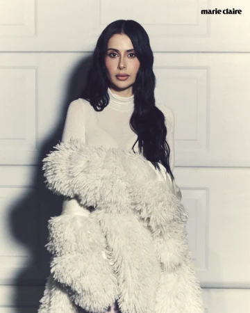 Portrait of a woman in a white turtleneck sweater and faux fur coat, posing indoors against a white paneled wall; Marie Claire logo visible.