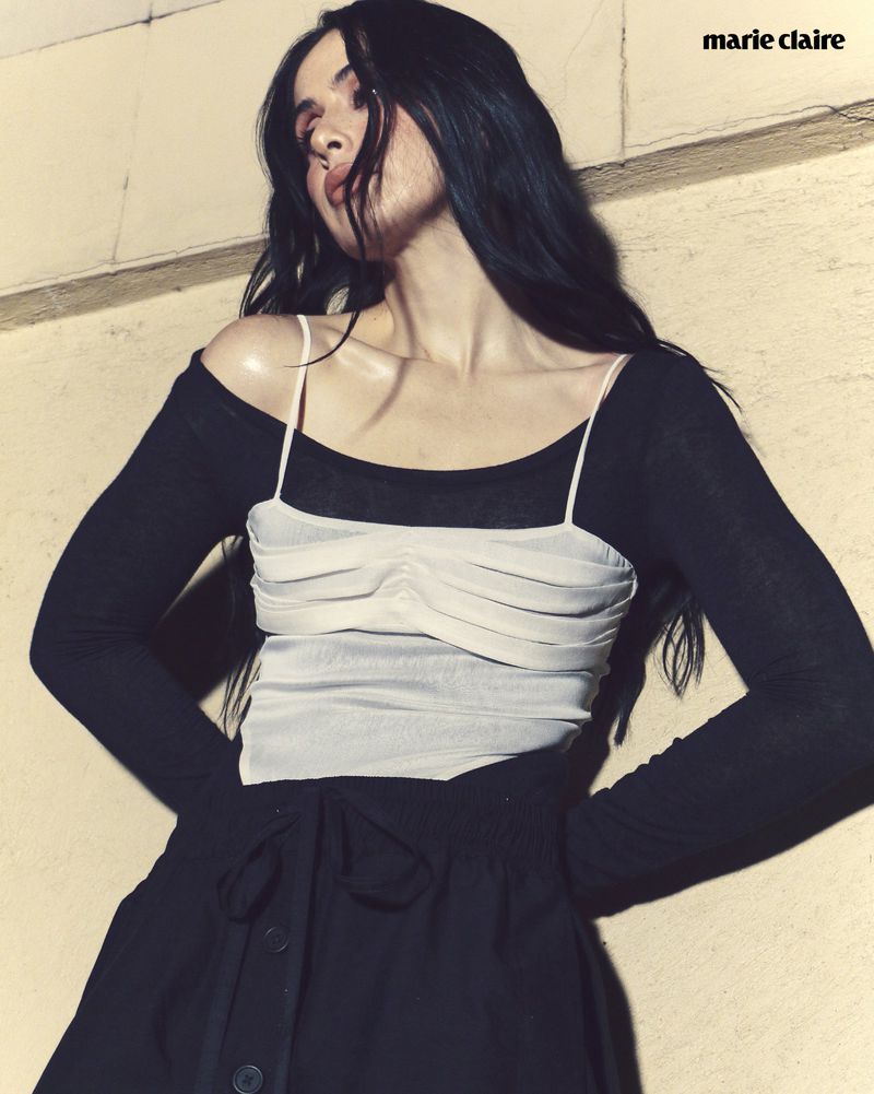 A person with long dark hair wearing a white top and black skirt is angled slightly looking up in an indoor setting with muted light.