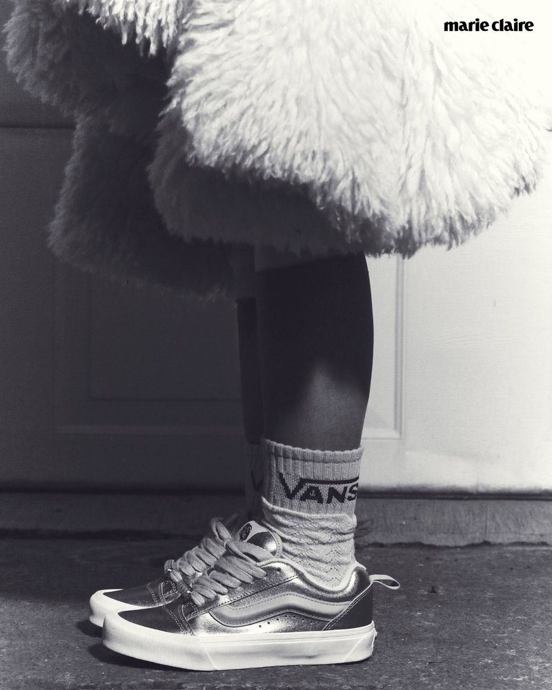 Close-up black and white shot of a person wearing metallic sneakers and socks, with part of a fluffy coat in the background.