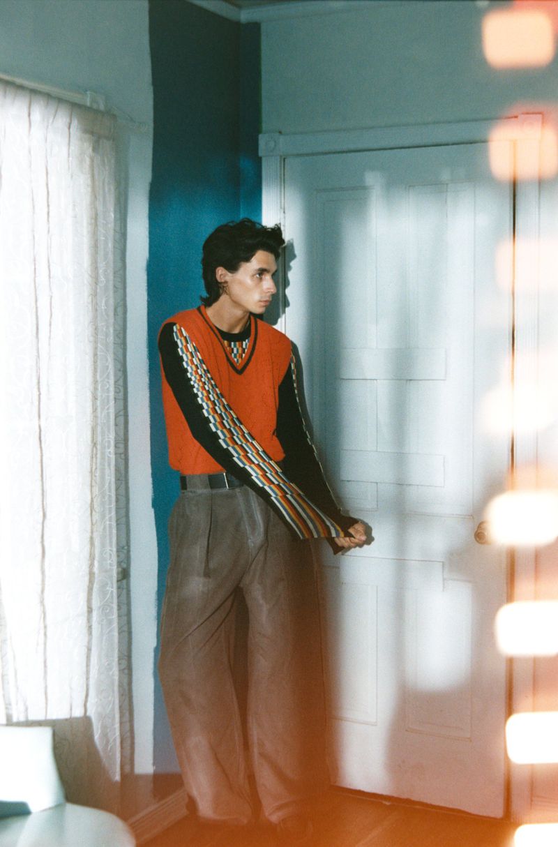 Young adult man in orange sweater vest stands by a white door, bathed in soft light, creating a casual and fashion-forward indoor scene.