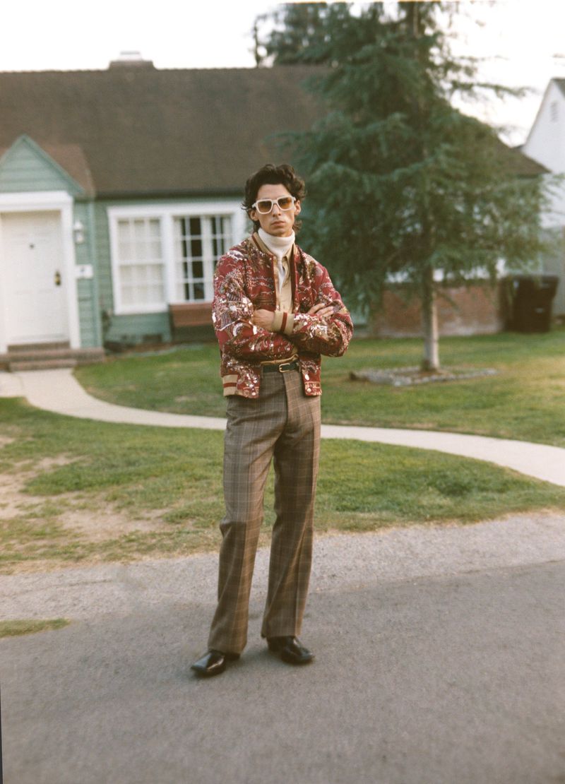 Adult male in printed bomber jacket, plaid pants, and sunglasses stands with arms crossed outside suburban house on sunny day.