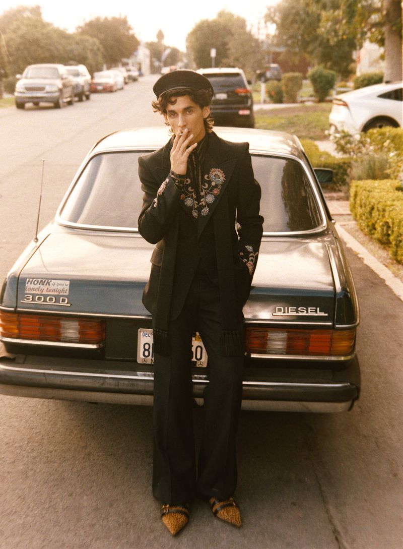 Male model in embroidered blazer and hat leans against classic car on street. Residential area background with cars and houses.