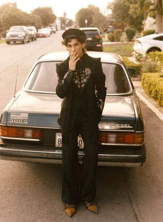 Male model in embroidered blazer and hat leans against classic car on street. Residential area background with cars and houses.