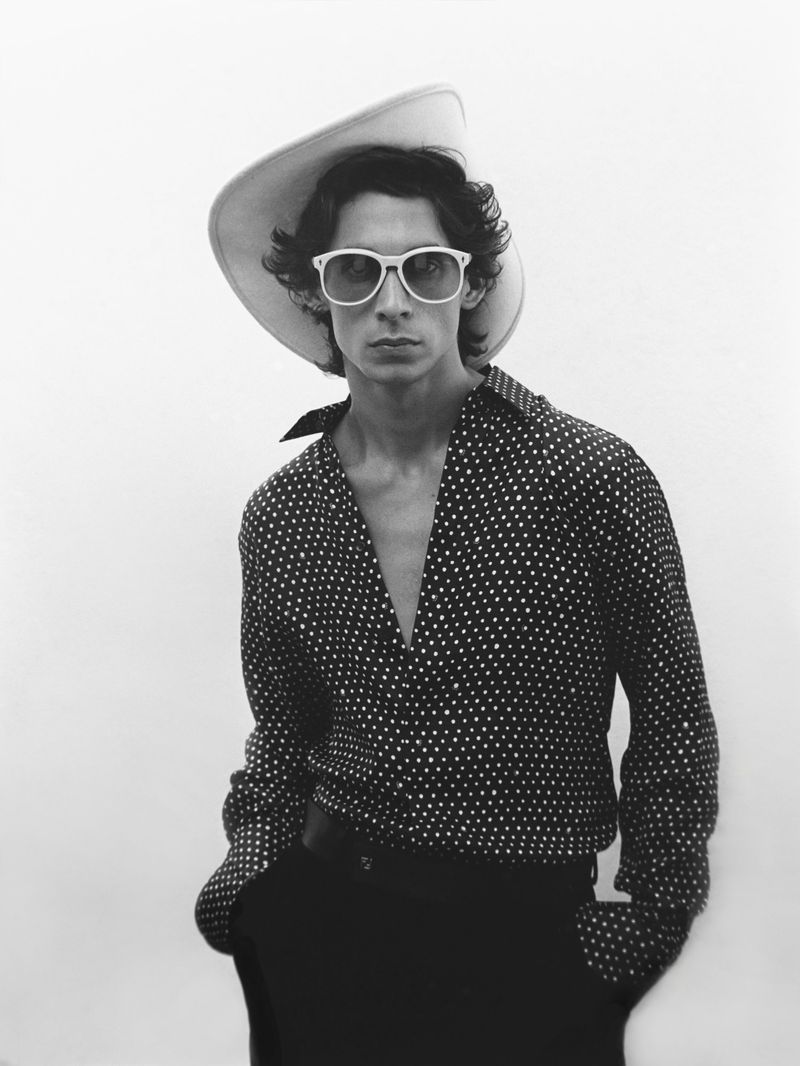 A black and white portrait of a slim adult male wearing a wide-brimmed hat, sunglasses, and a patterned shirt against a plain background.