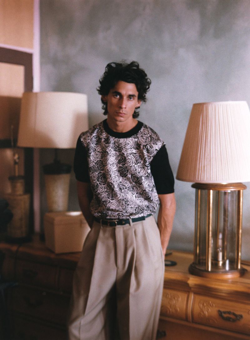 Portrait of a man with dark hair, wearing patterned shirt and tan pants, standing indoors with a neutral background, lamps visible.