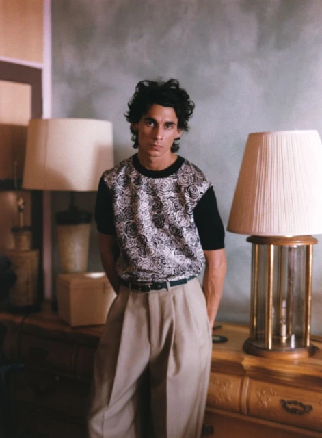 Portrait of a man with dark hair, wearing patterned shirt and tan pants, standing indoors with a neutral background, lamps visible.