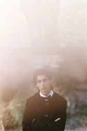Portrait of a man with arms crossed wearing a dark buttoned jacket and bow tie against a soft-focus outdoor background.