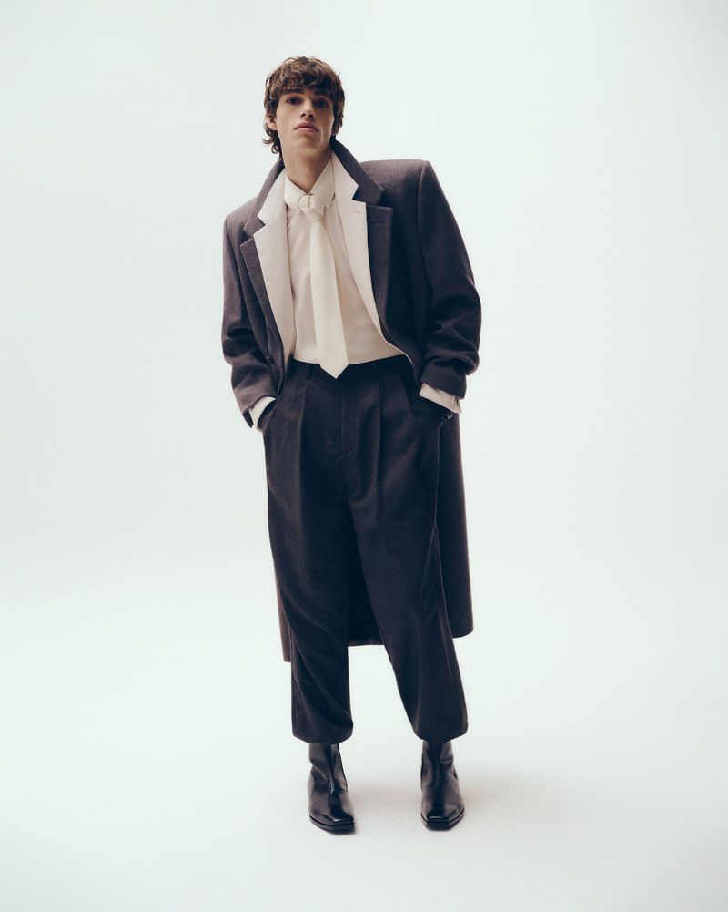 A full-length studio shot of a young adult male fashion model posing in a dark grey wool coat, baggy pleated trousers, and black Chelsea boots.