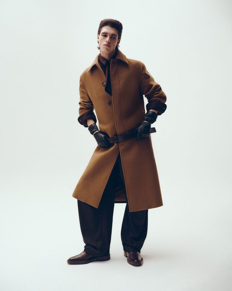 Full-length portrait of a male model wearing a camel coat and black gloves against a clean white studio background.