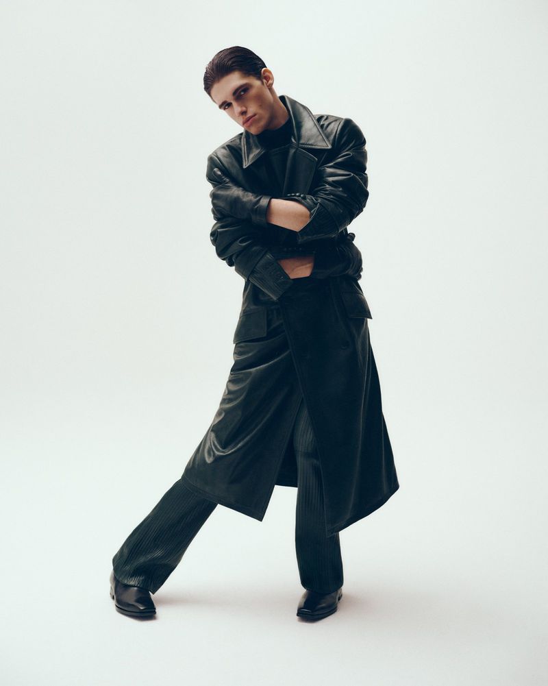 A stylish young adult male poses in a long black leather coat and trousers against a neutral backdrop in a fashion editorial-style photograph.