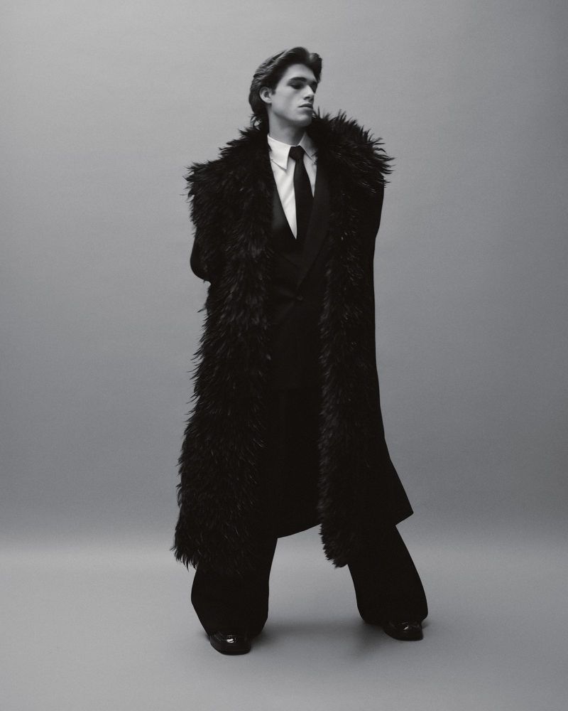 A man poses in a black suit and feathered coat in a studio setting. He has dark hair and is wearing a tie and dress shoes.