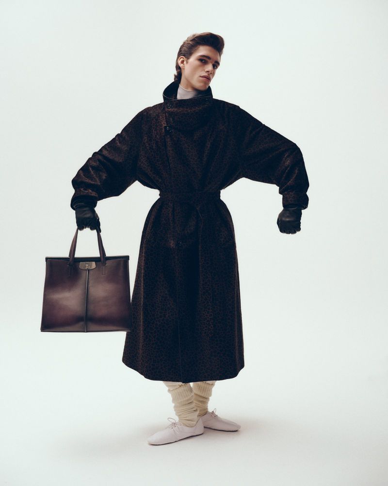 Model Theo Crown poses in an oversized dark belted overcoat holding a brown tote bag against a white studio background.