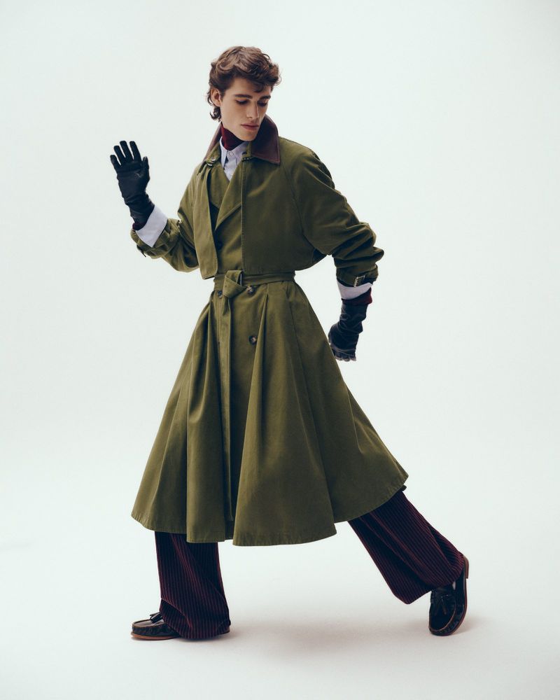 A stylish young adult man in an olive green trench coat, maroon pants, and black gloves waves in a studio fashion shot.