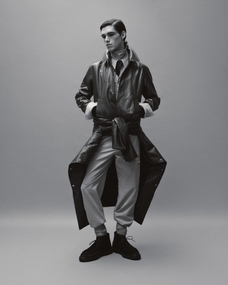 Monochrome fashion portrait of a male model in an open black trench coat, gray pants, and black boots against a gray backdrop.