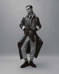 Model Theo Crown wearing a dark leather coat over light trousers and ankle boots against a grey studio backdrop.