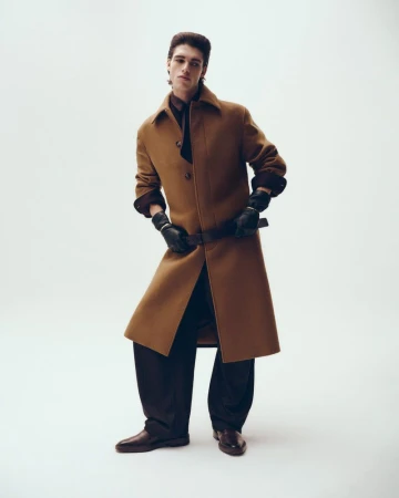 Full-length portrait of a male model wearing a camel coat and black gloves against a clean white studio background.