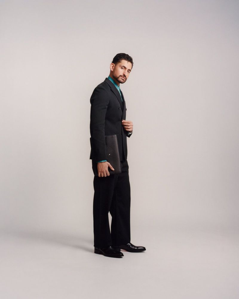 Full-length studio shot of adult male in a dark suit, holding a folder, gazing at the camera against a light-grey seamless backdrop.