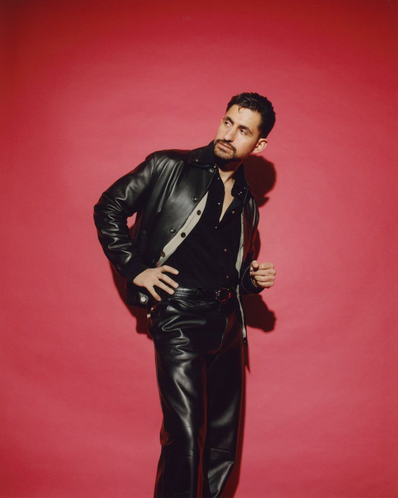 Stylish adult male in leather jacket and pants against a vibrant pink backdrop, looking up and to the side, capturing a fashionable pose.