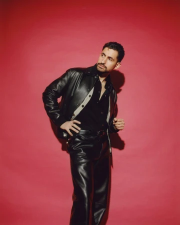 Stylish adult male in leather jacket and pants against a vibrant pink backdrop, looking up and to the side, capturing a fashionable pose.
