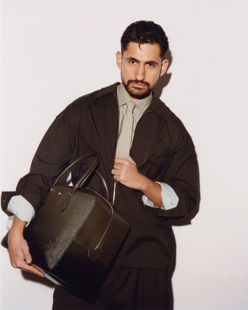 Adult man poses in a photo studio holding a dark-brown leather bag, wearing a brown jacket, light-green dress shirt, and a light-green tie.