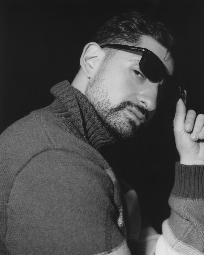 Black and white portrait of a bearded man wearing a ribbed knit turtleneck sweater and dark sunglasses against a black background.