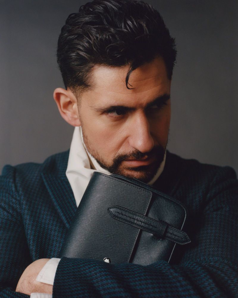 Man in blazer holds a black leather organizer against a gray background, looking toward the camera with a serious expression.