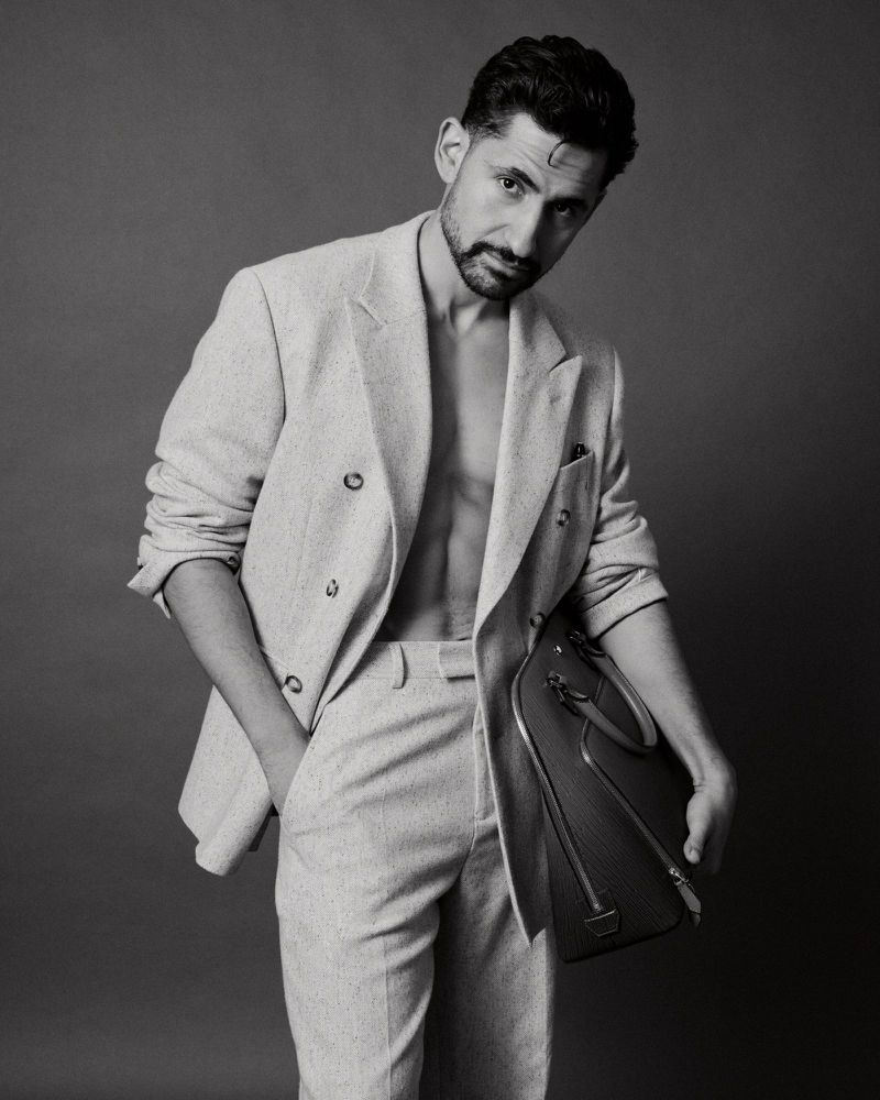 Monochrome studio portrait of a man in a suit, open jacket revealing torso, holding a structured leather bag, with a neutral background.