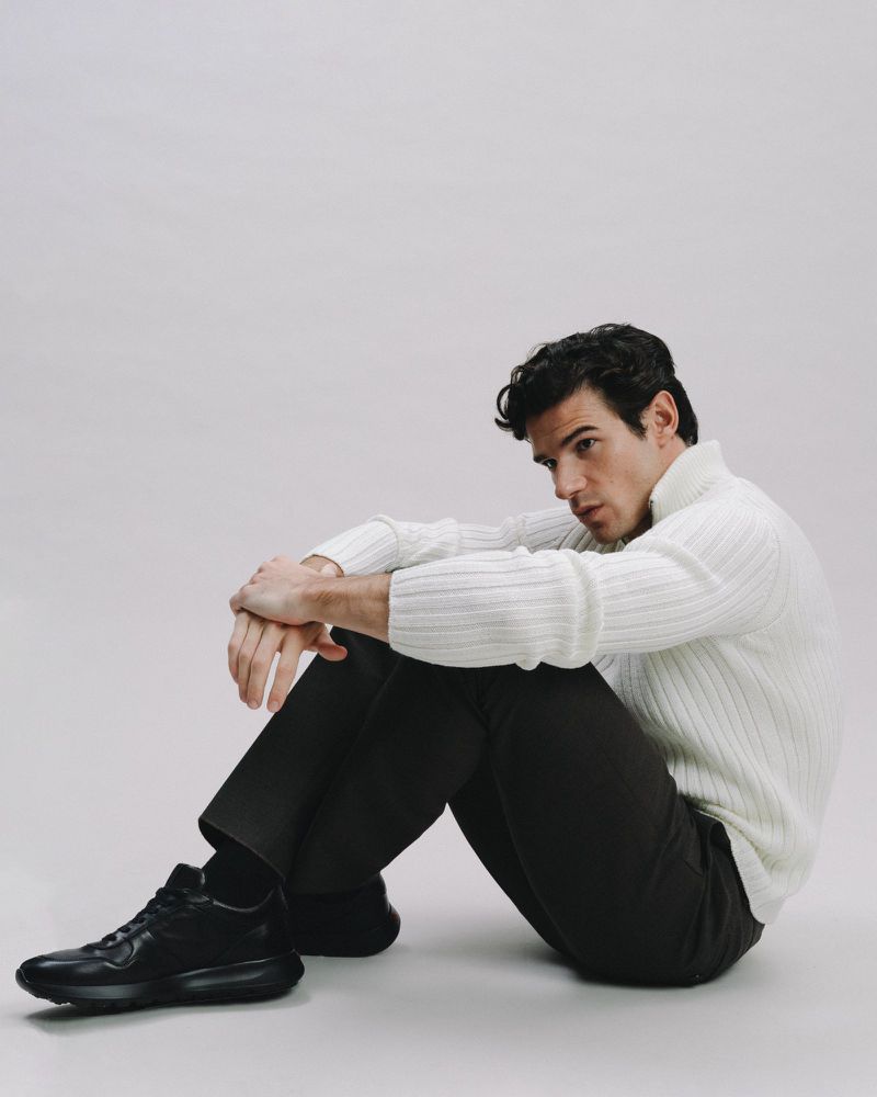 A young adult male sits in a white photo studio wearing a white sweater, dark pants, and black athletic shoes, looking to the side.