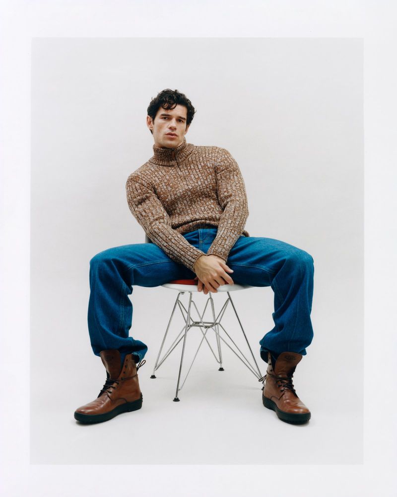 A young adult male sits on a white chair, wearing a brown turtleneck sweater, blue jeans, and brown boots against a white background.