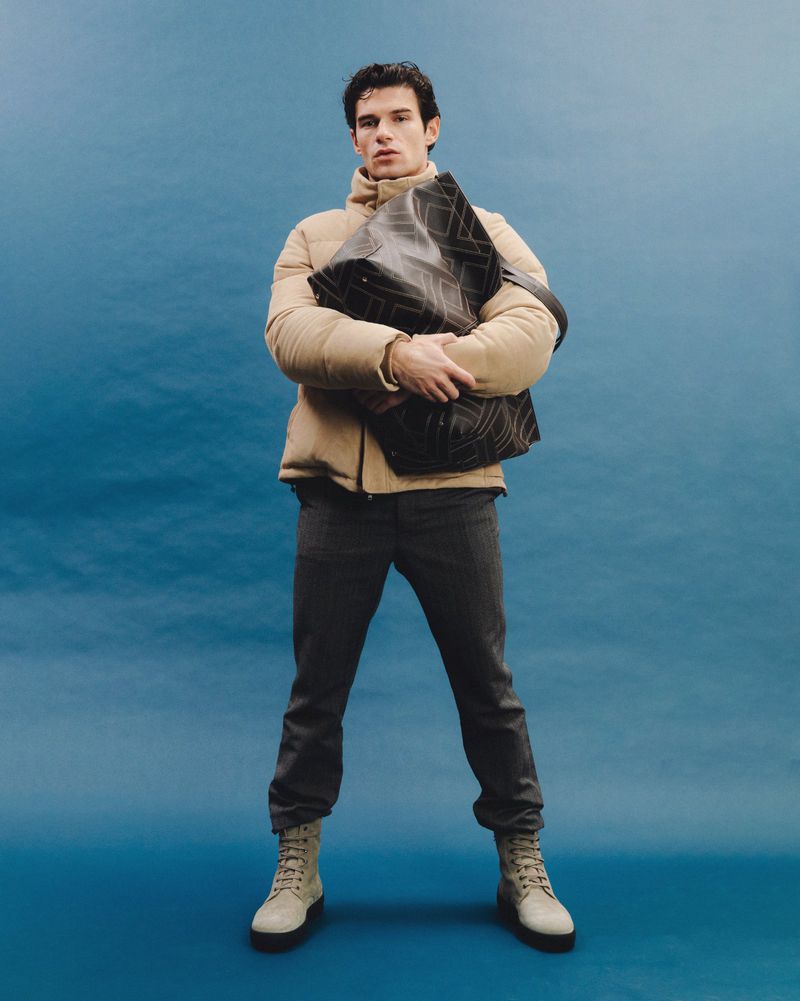 Full shot of a young adult male in a puffer jacket and dark jeans, holding a large black tote bag against a blue backdrop.
