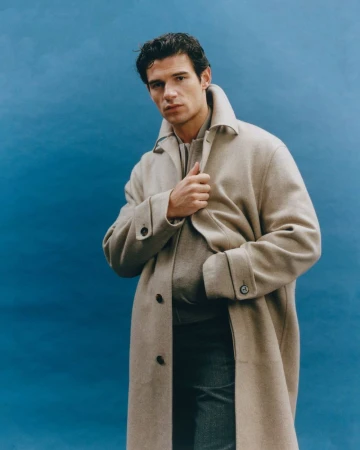 Man in a beige overcoat against a blue background looking at the camera. He is also wearing a sweater and jeans.