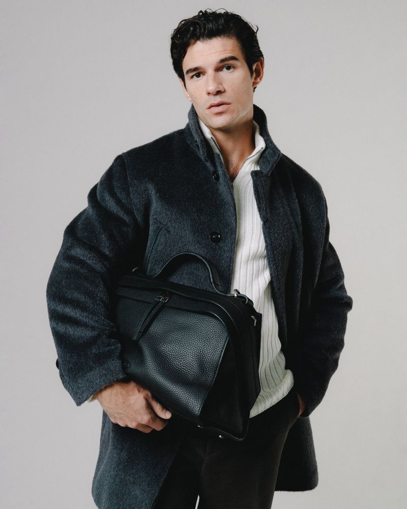 A man poses in a studio wearing a coat and sweater, holding a leather briefcase. He is facing forward with a neutral expression.